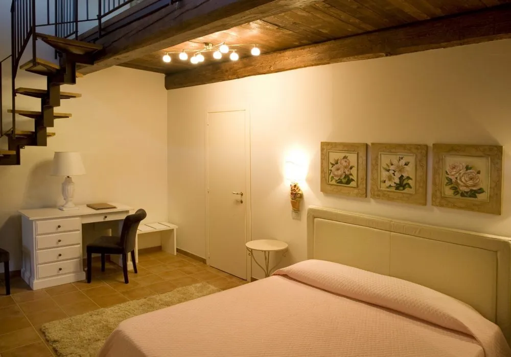 Camera in soppalco con letto dal copriletto rosa, tre quadretti floreali e scala a chiocciola in ferro