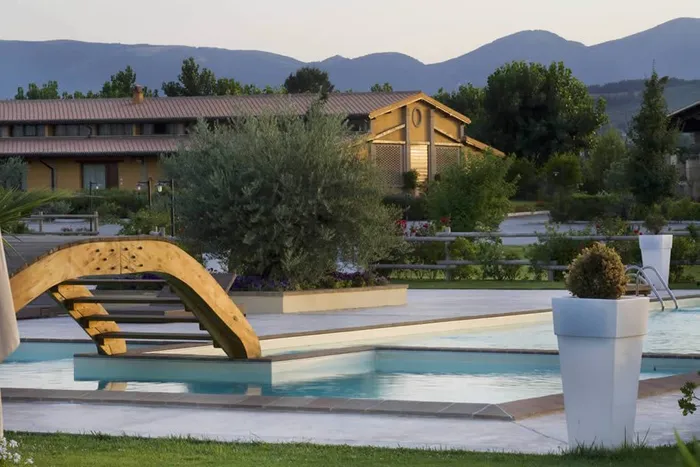 Piscina esterna al tramonto con ponticello in legno, ulivo e colline sullo sfondo